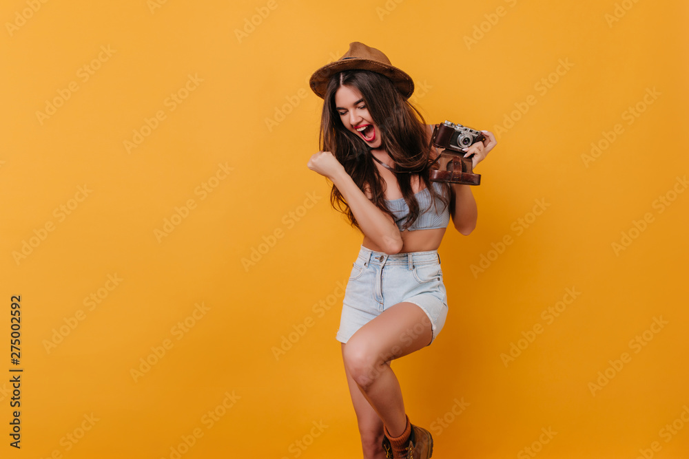Wonderful female photographer funny dancing on yellow background. Portrait of slim tanned girl with camera fooling aound in studio.