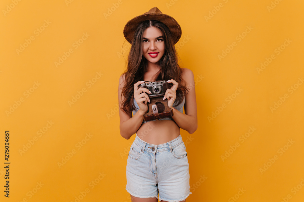 Shapely girl wears white shorts holding camera while standing on yellow background. Spectacular female photographer expressing positive emotions after photoshoot.
