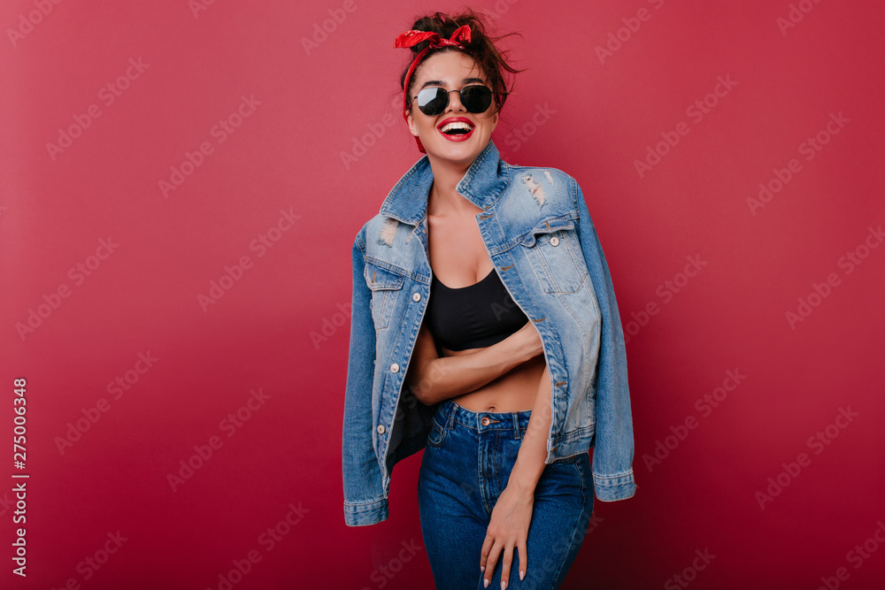 Pleased girl wears jeans laughing on claret background. Spectacular female model looking to camera through black sunglasses. - obrazy, fototapety, plakaty 