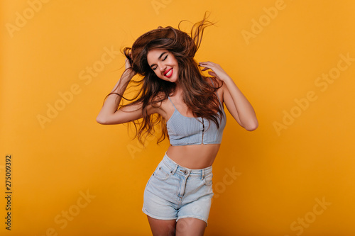 Ecstatic girl with inspired face expression dancing on yellow background. Studio shot of glamorous tanned female model in trendy shorts enjoying photoshoot.