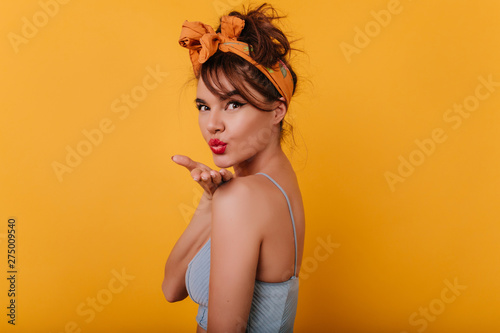 Beautiful brunette girl looking over shoulder and sending air kiss. Indoor portrait of relaxed tanned lady with orange ribbon in hair.
