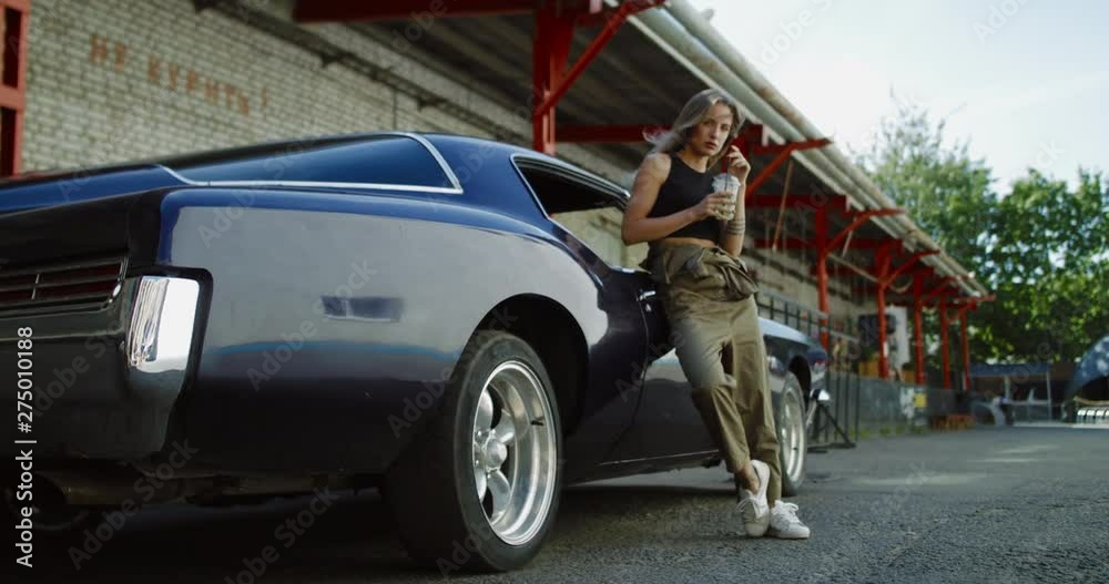 Beautiful Caucasian female posing with an old classic retro muscle car. Automotive lifestyle. 4K UHD RAW graded footage