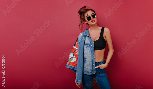Adorable tanned girl in black sunglasses standing in confident pose on claret background. Wonderful slim lady in sport tank-top posing in studio.