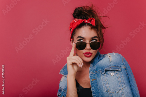 Adorable pensive girl in sunglasses posing on claret background. Graceful black-haired female model in vintage attire thinking about something.