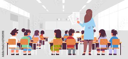 female teacher holding tests with good a grade rear view pupils group sitting in classroom during lesson teaching education concept modern class room interior horizontal full length