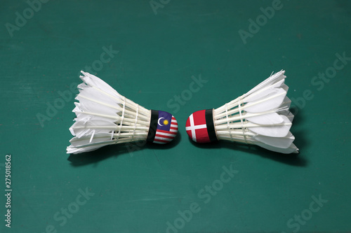 Two used shuttlecocks on green floor of Badminton court with both head each other. One head painted with Malaysia flag and one head painted with the Denmark flag.