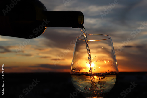Fototapeta Naklejka Na Ścianę i Meble -  White wine pouring from a bottle into the glass on beautiful sunset background, orange sun is shining through the jet. Concept of celebration, summer party at resort, romantic dinner outdoors