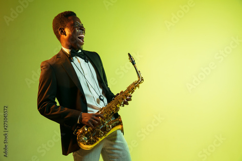 Young african-american jazz musician playing the saxophone on gradient yellow-green studio background. Concept of music  hobby  festival. Joyful attractive guy improvising. Colorful portrait of artist
