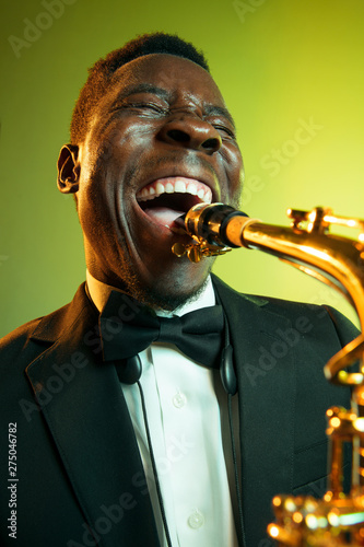 Canvas Print Young african-american jazz musician playing the saxophone on gradient yellow-green studio background