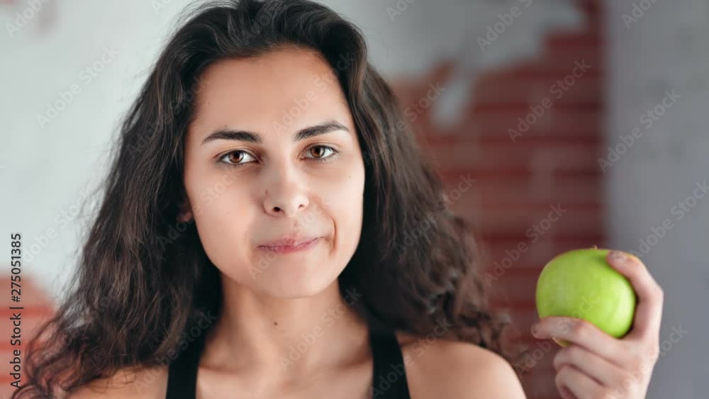 Close-up face of hungry beautiful young girl having perfect skin eating apple looking at camera