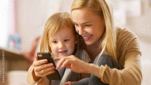 Close-up Shot of a Beautiful Young Mother with Her Little Daughter Playing with Smartphone. Mother Shows Her Something Interesting in It. Sunny Children's Room.