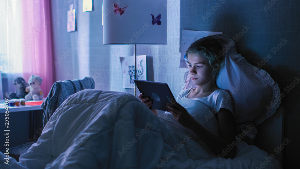 Fototapeta premium Young Teenage Girl Lies in Her Bed at Night With Tablet Computer. Watches TV Show and Smiles.