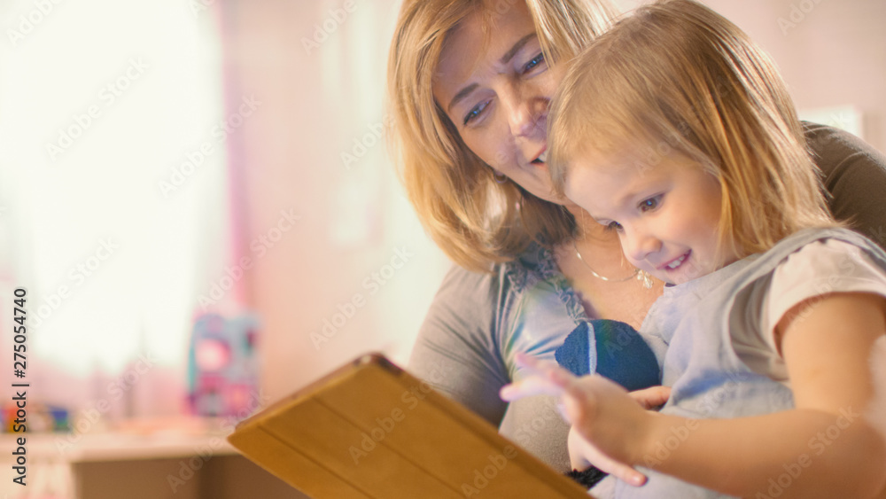 Beautiful Mother and Her Cute Little Daughter Sitting on the Floor and Reading Children's Books on a Tablet Computer. Sunny Living Room. Beautiful Mother and Her Cute Little Daughter Sitting on the Floor and Reading Children's Books on a Tablet Computer. Sunny Living Room.