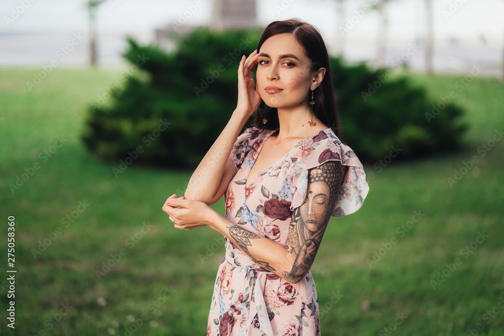 Girl with tattoo on her arm and in summer dress posing of greenery on background Girl with tattoo on her arm and in summer dress posing of greenery on background