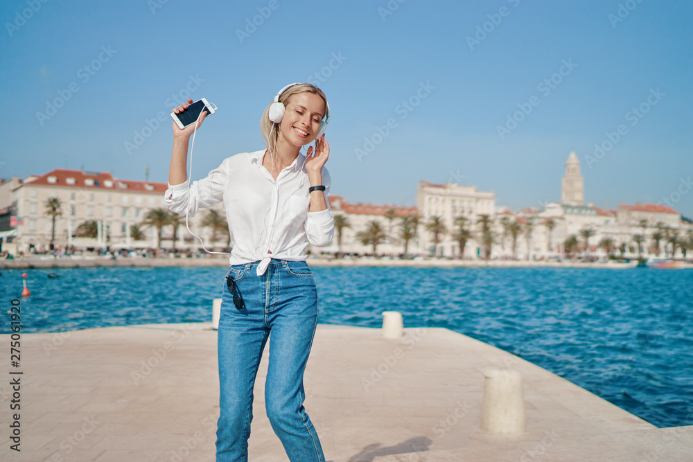 Holiday and music. Happy young woman with earphones and smartphone dancing on seafront promenade embankment.