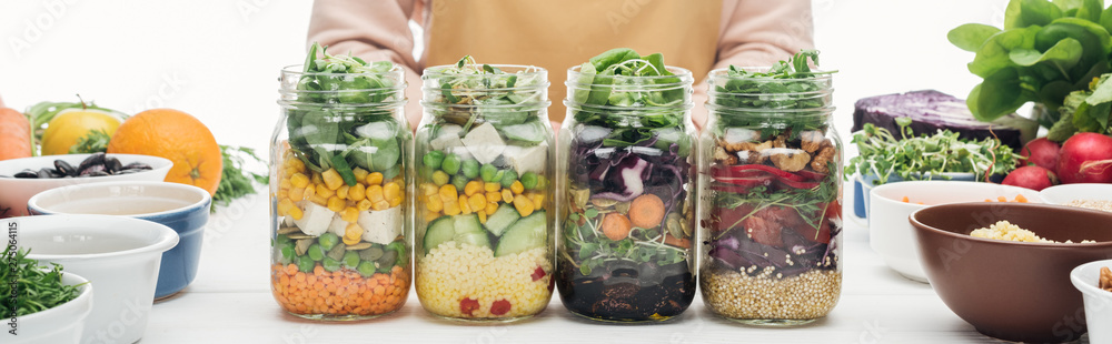 cropped view of woman in apron with glass jars with salad on wooden table isolated on white, panoramic shot cropped view of woman in apron with glass jars with salad on wooden table isolated on white, panoramic shot