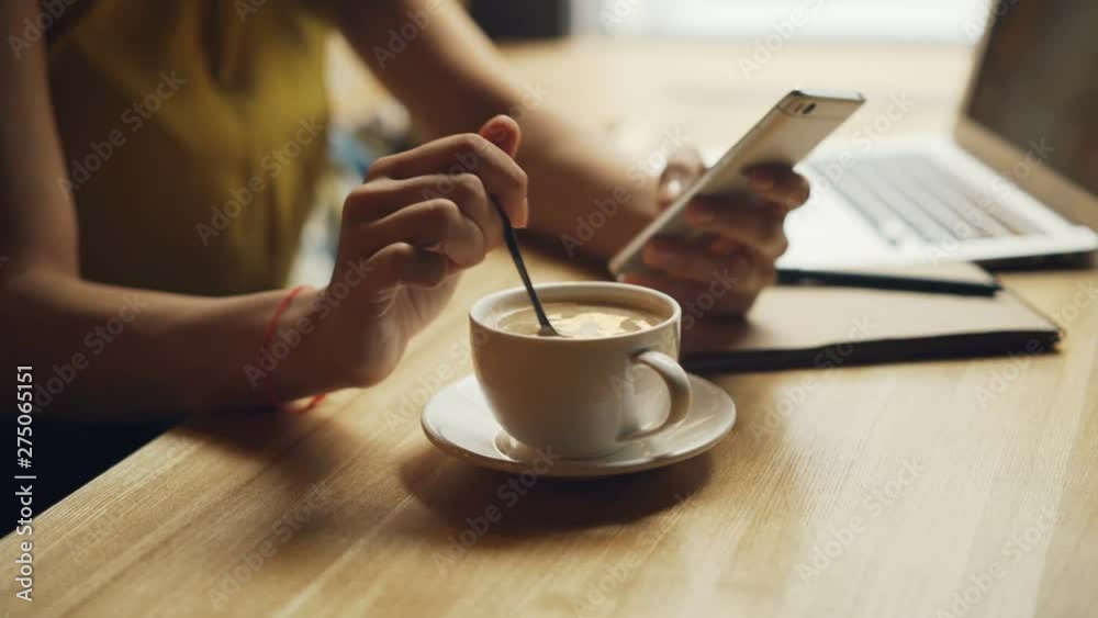 Close-up of woman's hands mixing coffee in cup and using smartphone relaxing in cafesitting at table alone. People, modern technology and drinks concept. Close-up of woman's hands mixing coffee in cup and using smartphone relaxing in cafesitting at table alone. People, modern technology and drinks concept.