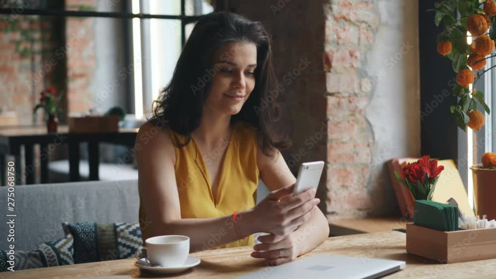 Charming young lady with black hair is holding smartphone touching screen smiling sitting at table in modern cafe. Cup of coffee and laptop are visible. Charming young lady with black hair is holding smartphone touching screen smiling sitting at table in modern cafe. Cup of coffee and laptop are visible.