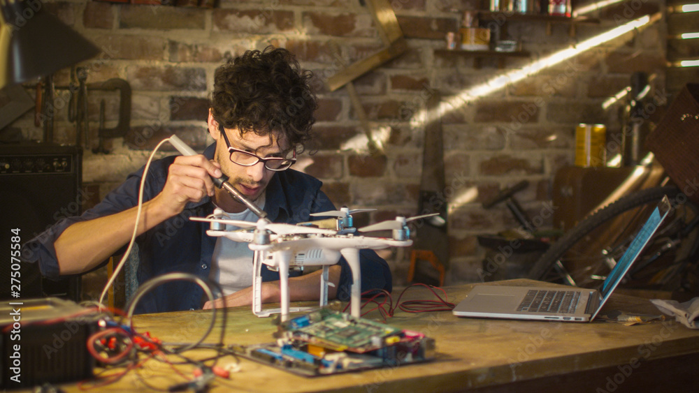 Man is soldering electrical components on a drone in a garage while checking a laptop computer. Man is soldering electrical components on a drone in a garage while checking a laptop computer.