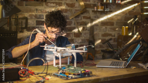 Man is soldering electrical components on a drone in a garage while checking a laptop computer.