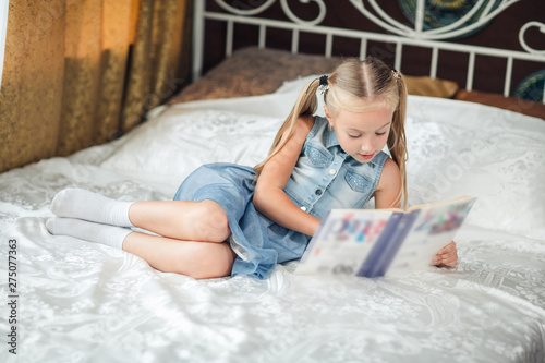 Home portrait of a little girl in denim sundress with long blond hair sitting in a bed and reading a book