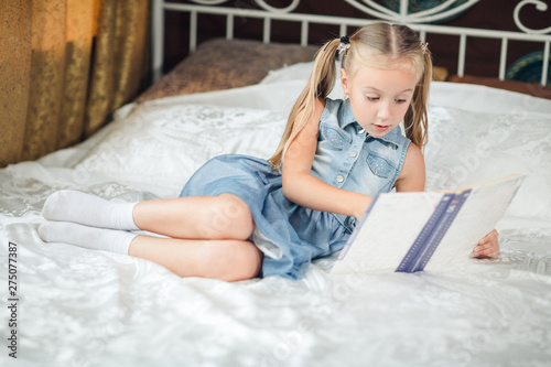 Cute little girl in denim sundress reading book on bed at home