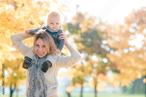 Young mother posing with little child on her shoulders in sunny autumn park.