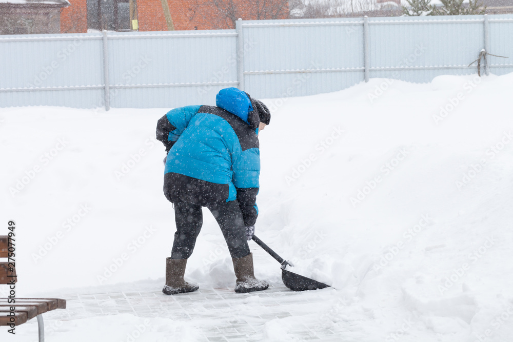 Old woman in warm blue jacket clears a snowdrifts with a snow shovel