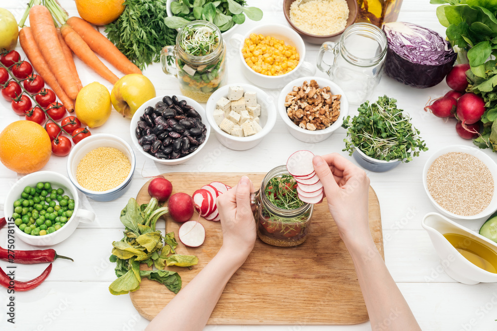 cropped view of woman adding radish slices in glass jar on wooden white table