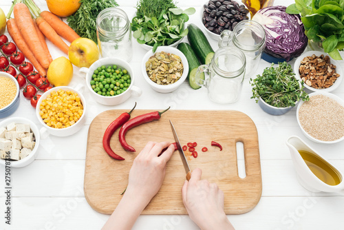 cropped view of woman cutting chili peppers on wooden chopping board on white table