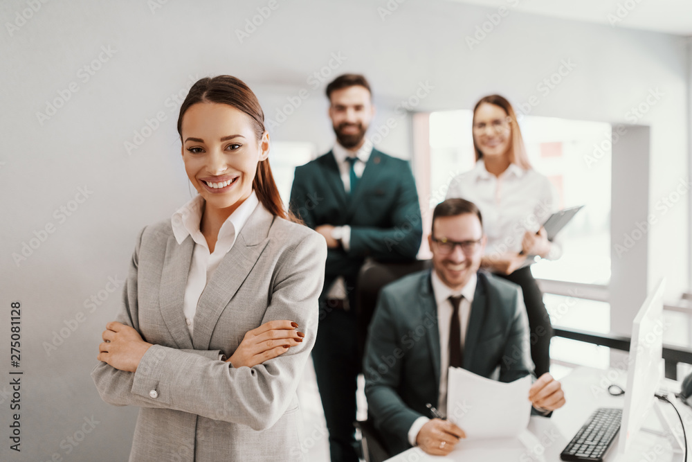 Gorgeous smiling businesswoman in suit standing in office with hands crossed. In background her successful team posing. Gorgeous smiling businesswoman in suit standing in office with hands crossed. In background her successful team posing.