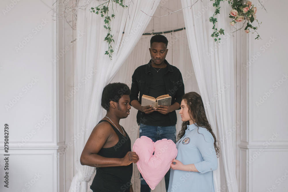 Fotka „Interracial couple standing on knees in front of wedding arch. Interracial bridal ceremony. Caucasian woman getting married with her african dark skinned boyfriend. Indoor amateur wedding ceremony.“ ze služby Stock |