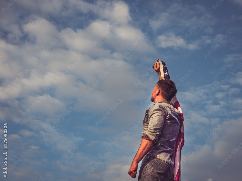 Attractive man in jeans and denim shirt waving an American Flag against a clear, sunny, blue sky. View from the back, close-up. National holiday concept Attractive man in jeans and denim shirt waving an American Flag against a clear, sunny, blue sky. View from the back, close-up. National holiday concept