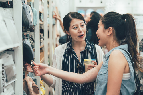 Two smiling female friends selecting products in grocery store. young girls discussing about summer trip to usa buying pillow bag on shelf hold look. happy asian women talking laugh in shop hong kong photo