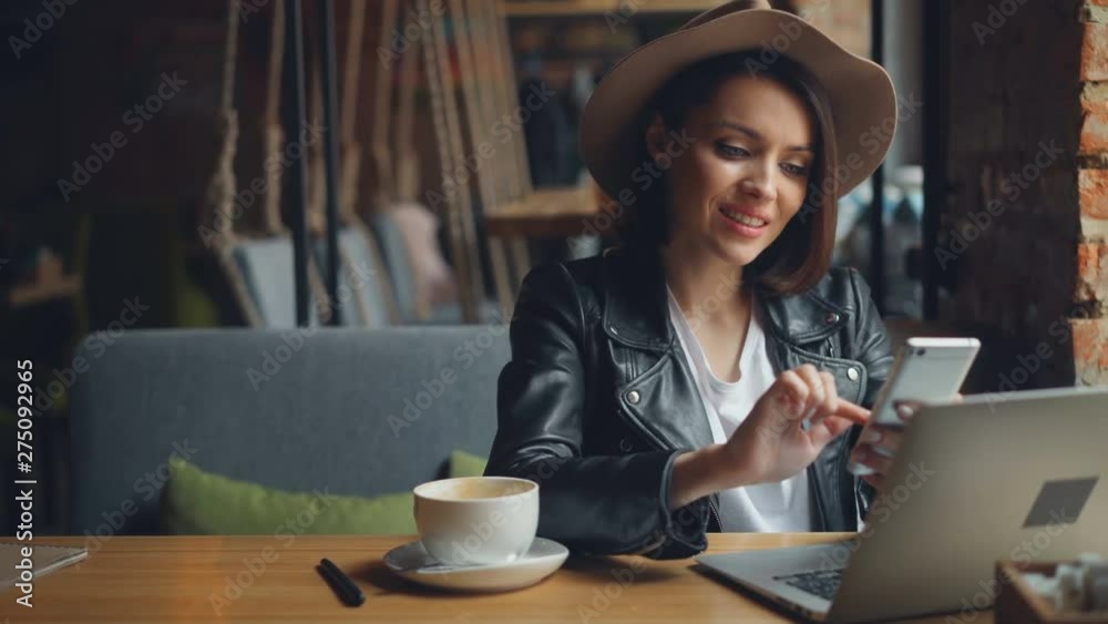 Attractive young woman hipster is using smartphone in cafe enjoying social media smiling sitting at table relaxing. Modern lifestyle and device concept.