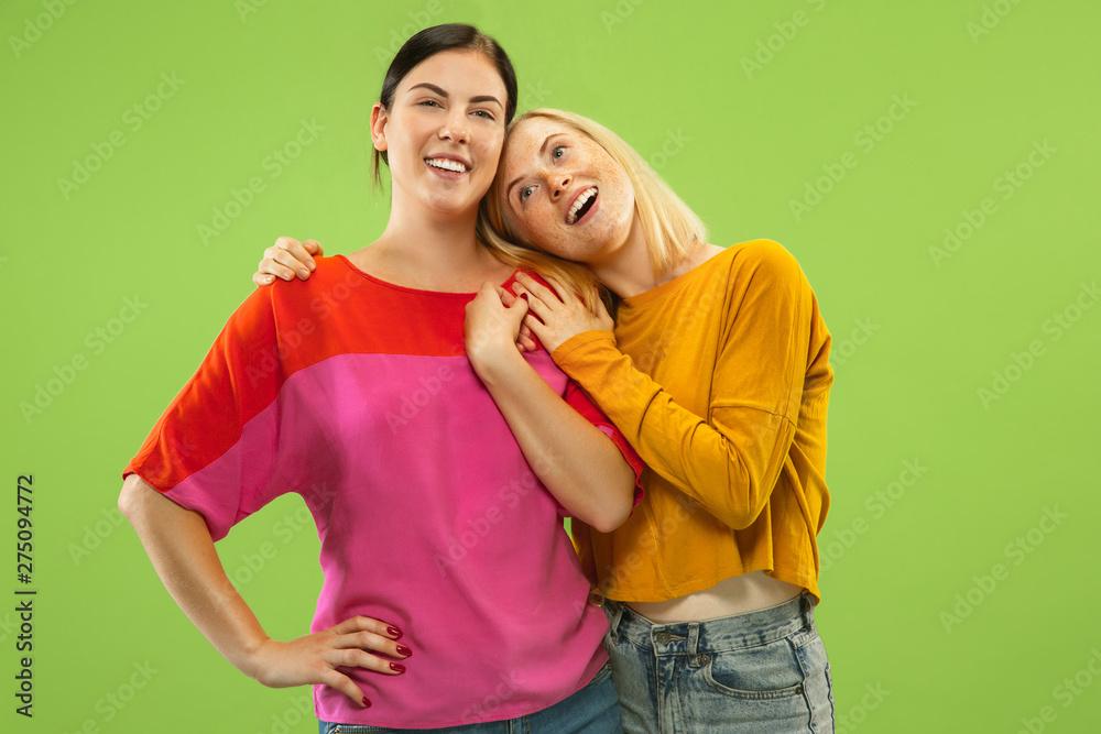 Naklejka premium Portrait of pretty charming girls in casual outfits isolated on green studio background. Two female models as a girlfriends or lesbians. Concept of LGBT, equality, human emotions, love, relation.