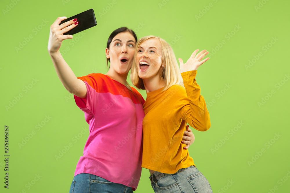 Portrait of pretty charming girls in casual outfits isolated on green studio background. Girlfriends or lesbians making selfie. Concept of LGBT, equality, human emotions, love, relation. Portrait of pretty charming girls in casual outfits isolated on green studio background. Girlfriends or lesbians making selfie. Concept of LGBT, equality, human emotions, love, relation.
