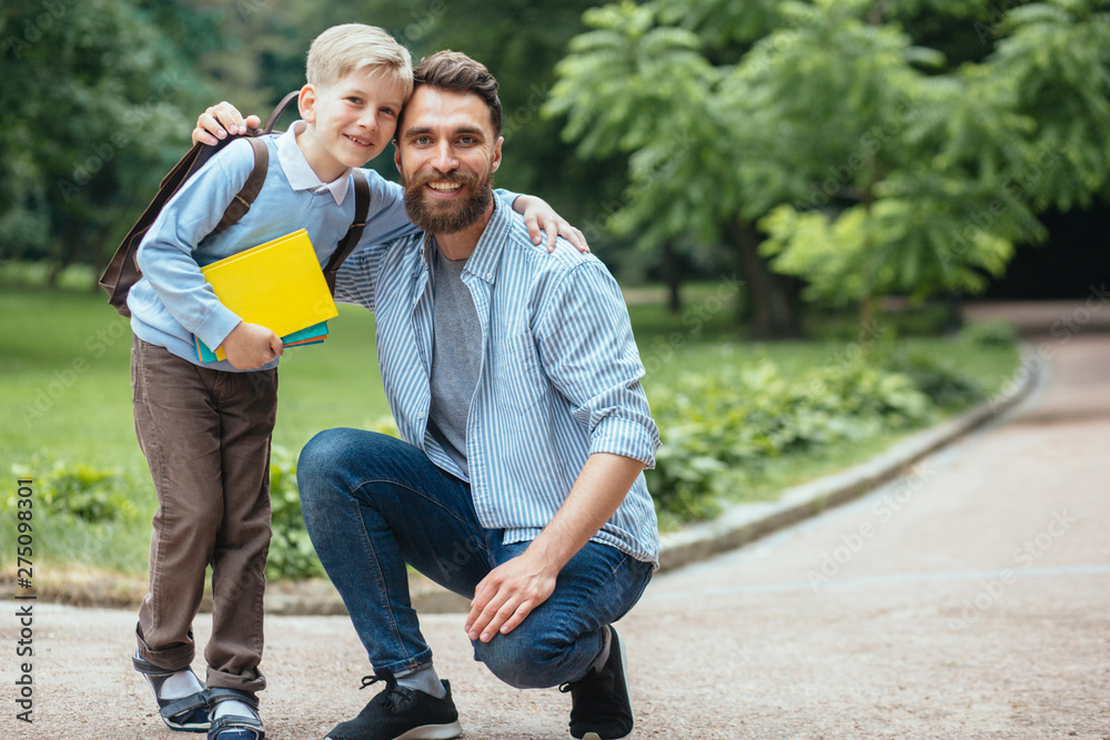 Back to school. Portrait of father and son looking at camera and smiling with park on background. They are go to school. Parent taking child to school. Pupil go study with backpack. First day of fall.