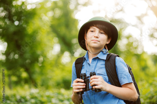 Portrait of little explorer with binoculars in forest. Boy traveler in helmet play in the park. Happy child go hiking with backpack in summer nature.