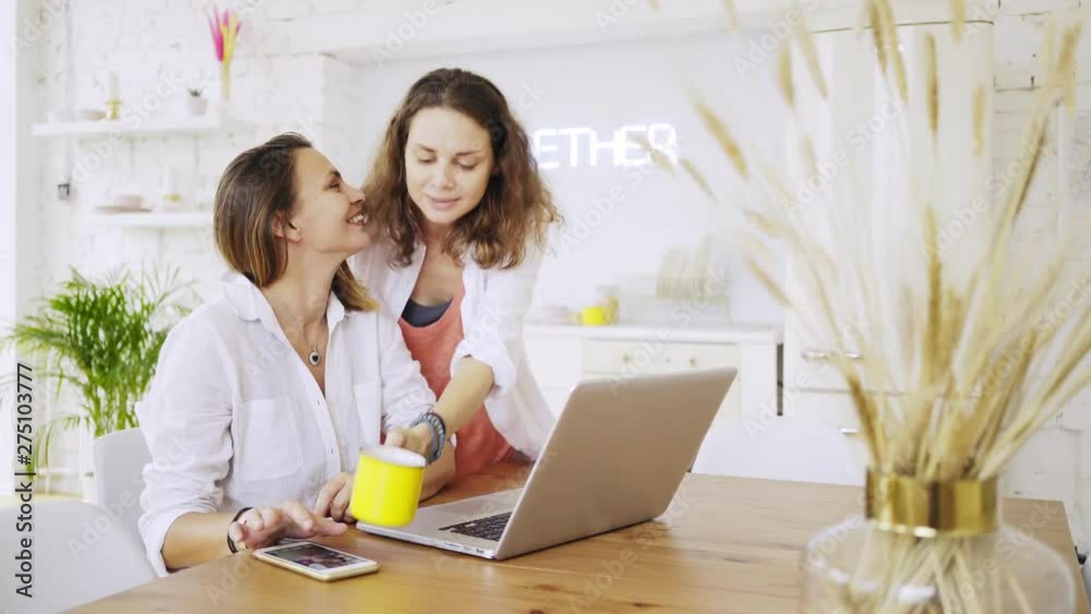 Morning in modern apartment . One girl working on laptop at the kitchen table, her girlfriend comes with breakfast, kisses on the cheek and together watching at display. Concept LGBT couple at home Morning in modern apartment . One girl working on laptop at the kitchen table, her girlfriend comes with breakfast, kisses on the cheek and together watching at display. Concept LGBT couple at home