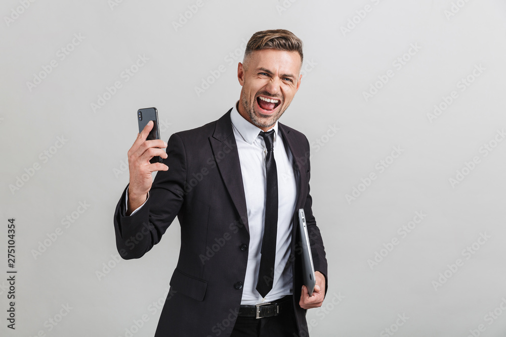 Naklejka premium Portrait of ecstatic adult businessman in office suit holding smartphone and laptop while standing