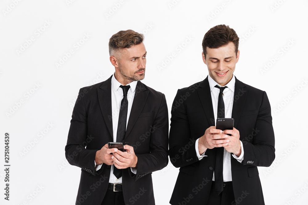 Image of handsome caucasian two businessmen in office suits holding and typing on cellphones Image of handsome caucasian two businessmen in office suits holding and typing on cellphones