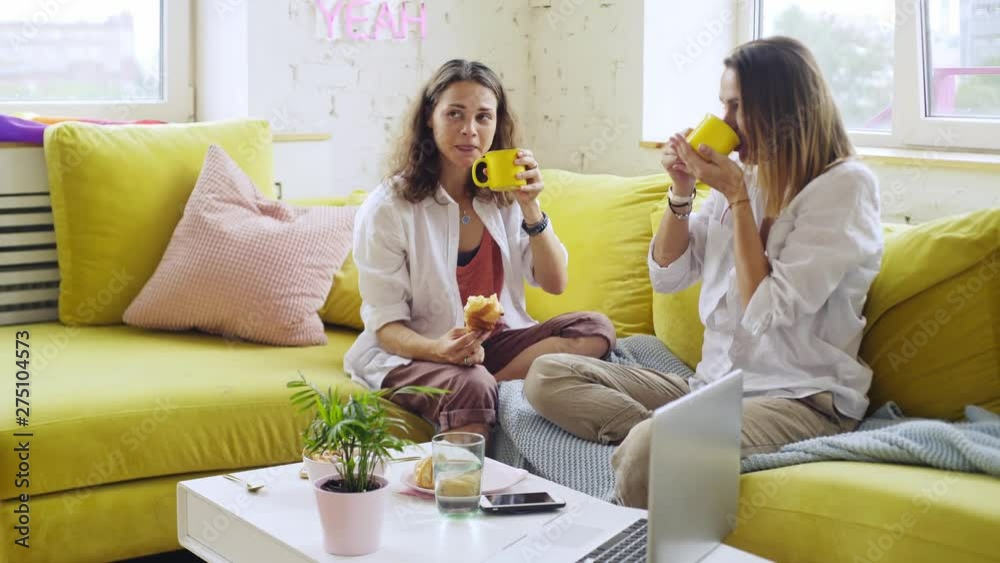 Girlfriends sitting on colorful sofa in modern apartment. Girls have breakfast, holding cup of coffee and eating croissant. Couple happily smiling enjoying togetherness. Concept LGBT couple at home Girlfriends sitting on colorful sofa in modern apartment. Girls have breakfast, holding cup of coffee and eating croissant. Couple happily smiling enjoying togetherness. Concept LGBT couple at home