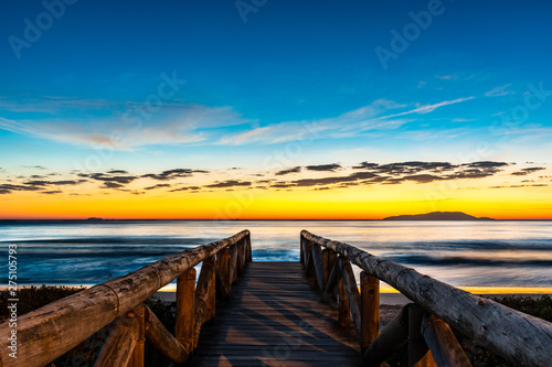 landscape with wooden walkway to seascape beach at sunrise with blue and orange sky and island in the background