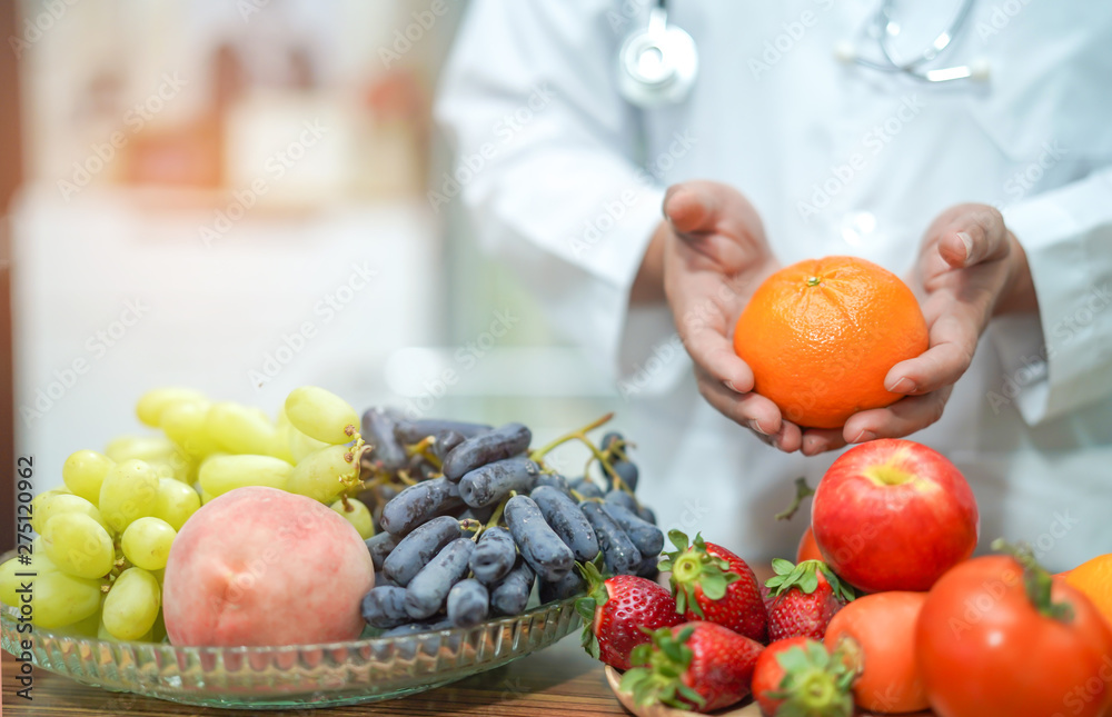 Nutritionist doctor holding orange juice with strawberry, potatoes, apples, plum, grapes, carrot, peach, pumpkin and salad vegetable on desk to instruction healthy food strong patient.