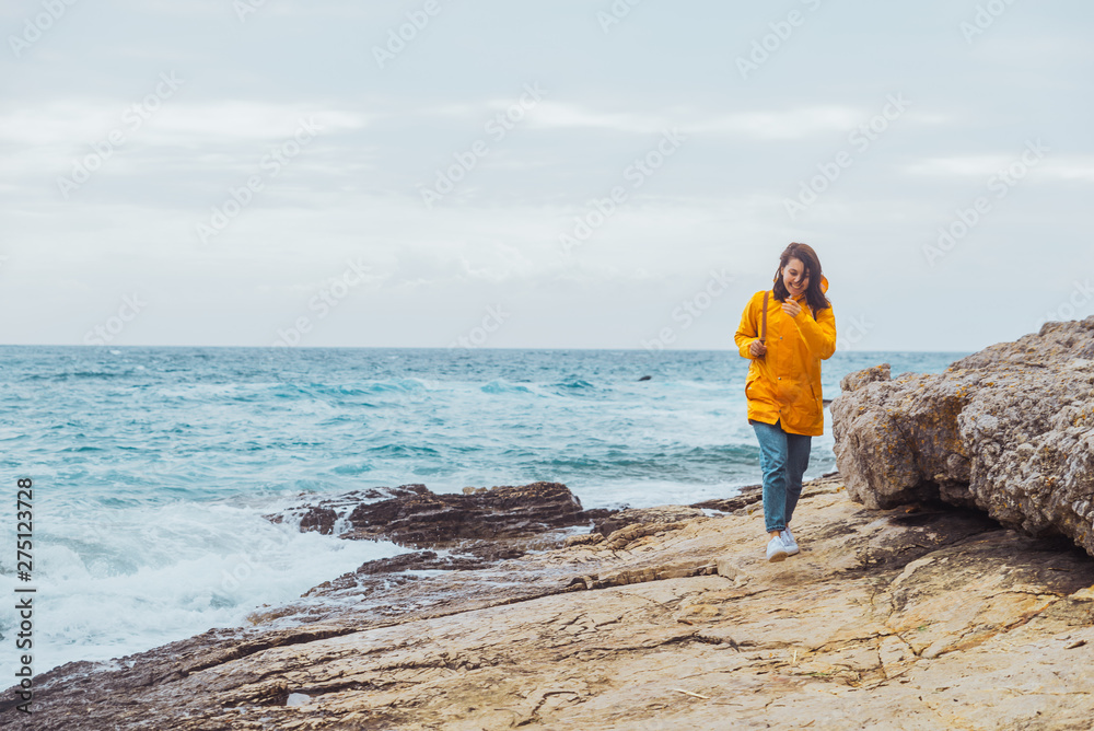 smiling woman in yellow raincoat walking by rocky beach stormy sea on background