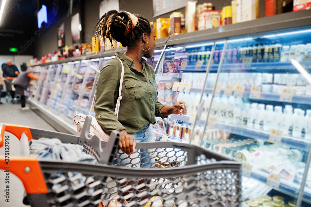 African woman with shopping cart choose yogurt bottle from fridge at supermarket.