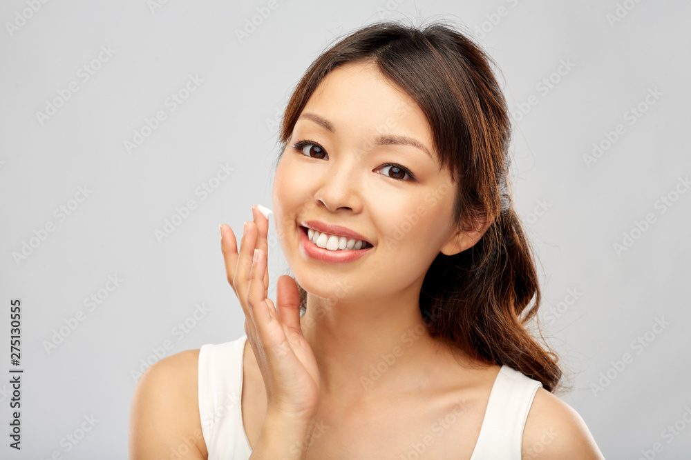 beauty, cosmetics and skincare concept - happy young asian woman applying moisturizer cream to her face over grey background beauty, cosmetics and skincare concept - happy young asian woman applying moisturizer cream to her face over grey background