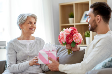 family, mother's day and birthday concept - smiling adult son giving present and flowers to his senior mother at home