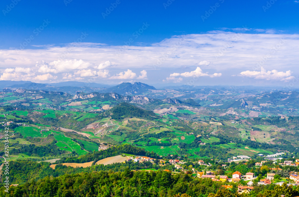 Aerial top panoramic view of landscape with valley, green hills, fields and villages of Republic San Marino suburban district with blue sky white clouds background. View from San Marino fortress